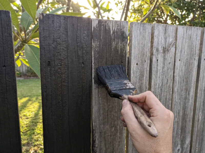 A brush applying charcoal stain to a timber paling fence, half coated and half weathered